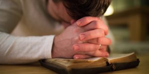 Unrecognizable young man praying, kneeling on the floor, hands on his Bible. Close up.